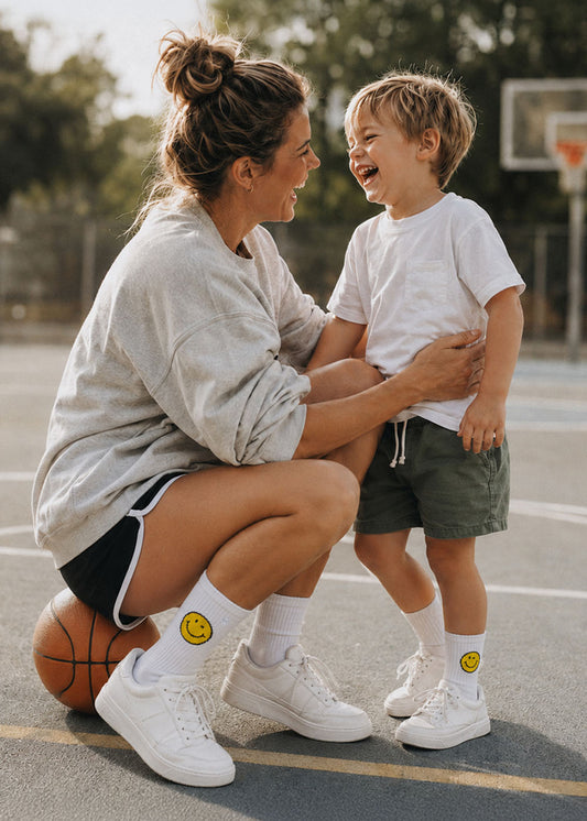 Eine lächelnde Frau und ein Junge tragen das „J.Clay Yellow Smile – Fam Pack“ auf einem Basketballplatz im Freien.