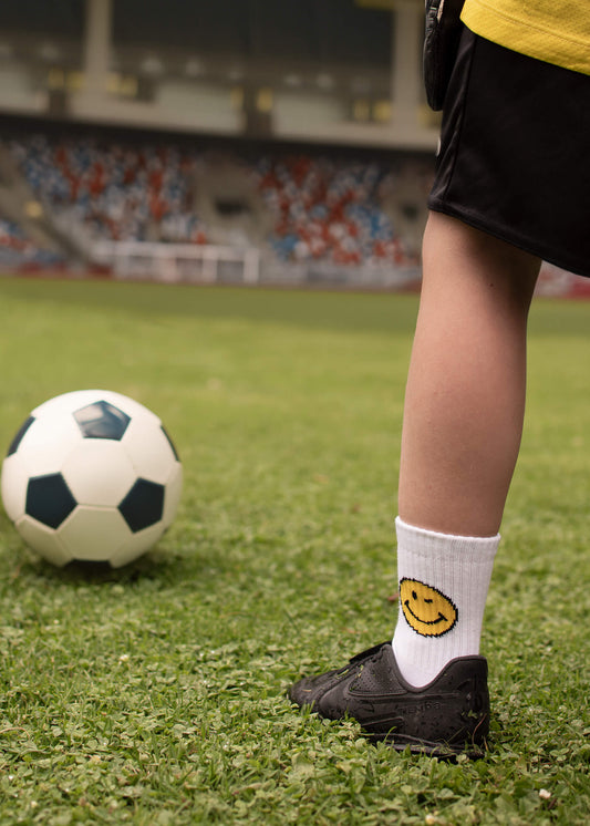 Eine Person in schwarzen Shorts, schwarzen Schuhen und J.Clay Kinder Tennissocken mit einem gelben Smiley-Gesicht steht auf einem Fußballfeld neben einem Ball und Stadionsitzen. Diese Kindersocken ab 5 Jahren von J.Clay sind ideal für aktive Kinder!.