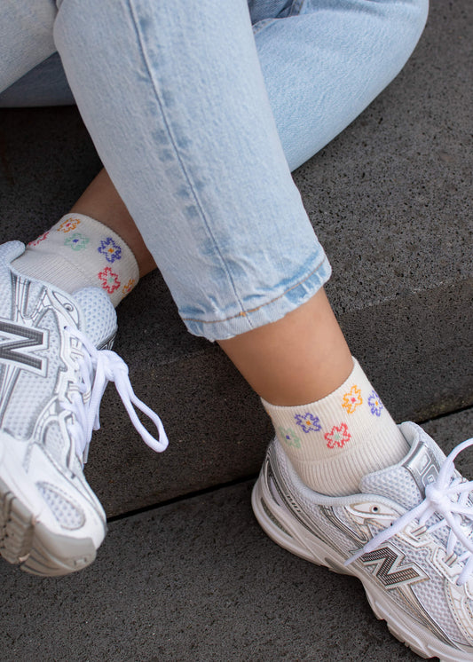 Eine Person in hellblauen Jeans, weißen Turnschuhen und J.Clay Sneaker Socken aus Baumwollmix mit farbenfrohem Blumenmuster sitzt auf einer grauen Betonfläche.