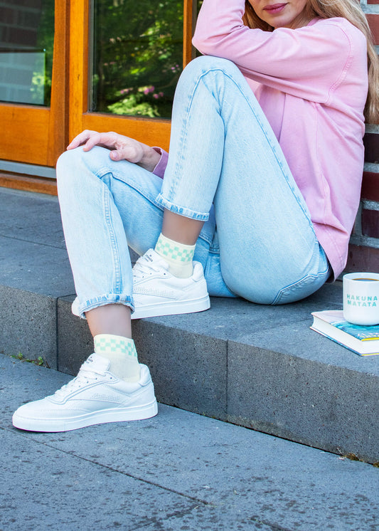 Eine Person in einem rosa Pullover und hellblauen Jeans sitzt auf einer Treppe im Freien und trägt J.Clay Sneaker Socken mit weißen Turnschuhen. In der Nähe steht eine Tasse mit der Aufschrift "Hakuna Matata". Ihr Gesicht ist teilweise außerhalb des Rahmens.