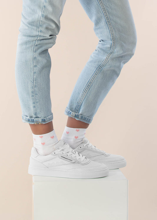 Eine Person in hellblauen, hochgekrempelten Jeans, weißen Turnschuhen und J.Clay Sneaker Socken mit rosa Herzmustern steht auf einem weißen Podest vor einem hellen Hintergrund.