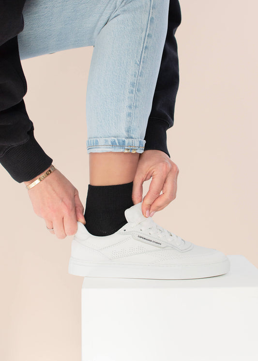 Eine Person in hellblauen Jeans und einem schwarzen Pullover passt die Schnürsenkel von weißen J.Clay Sneakers an, während sie auf einer weißen Fläche steht. Der Look wird mit schwarzen Socken und einem goldenen Armband abgerundet.