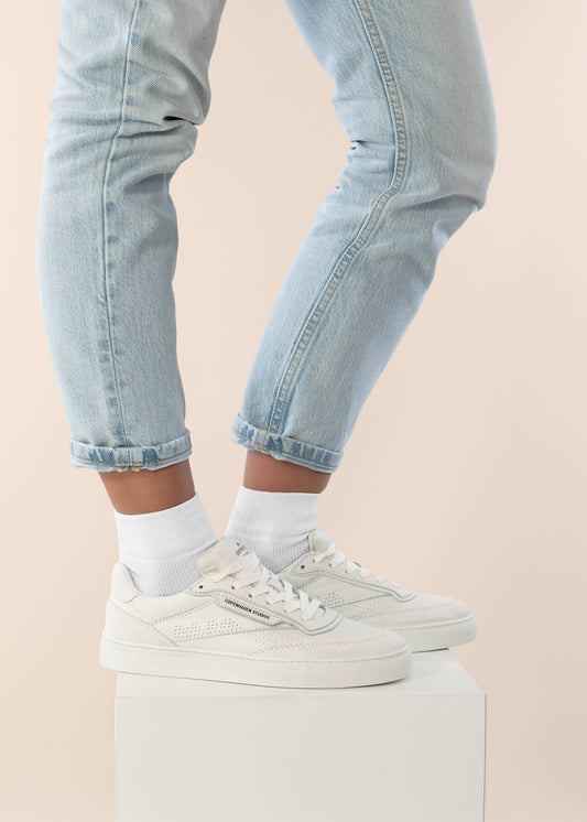 Eine Person, die J.Clay Sneaker Socken, hellblaue Jeans und weiße Turnschuhe trägt, steht auf einem weißen Block, wobei nur ihre Beine und Füße vor einem hellbeigen Hintergrund zu sehen sind.