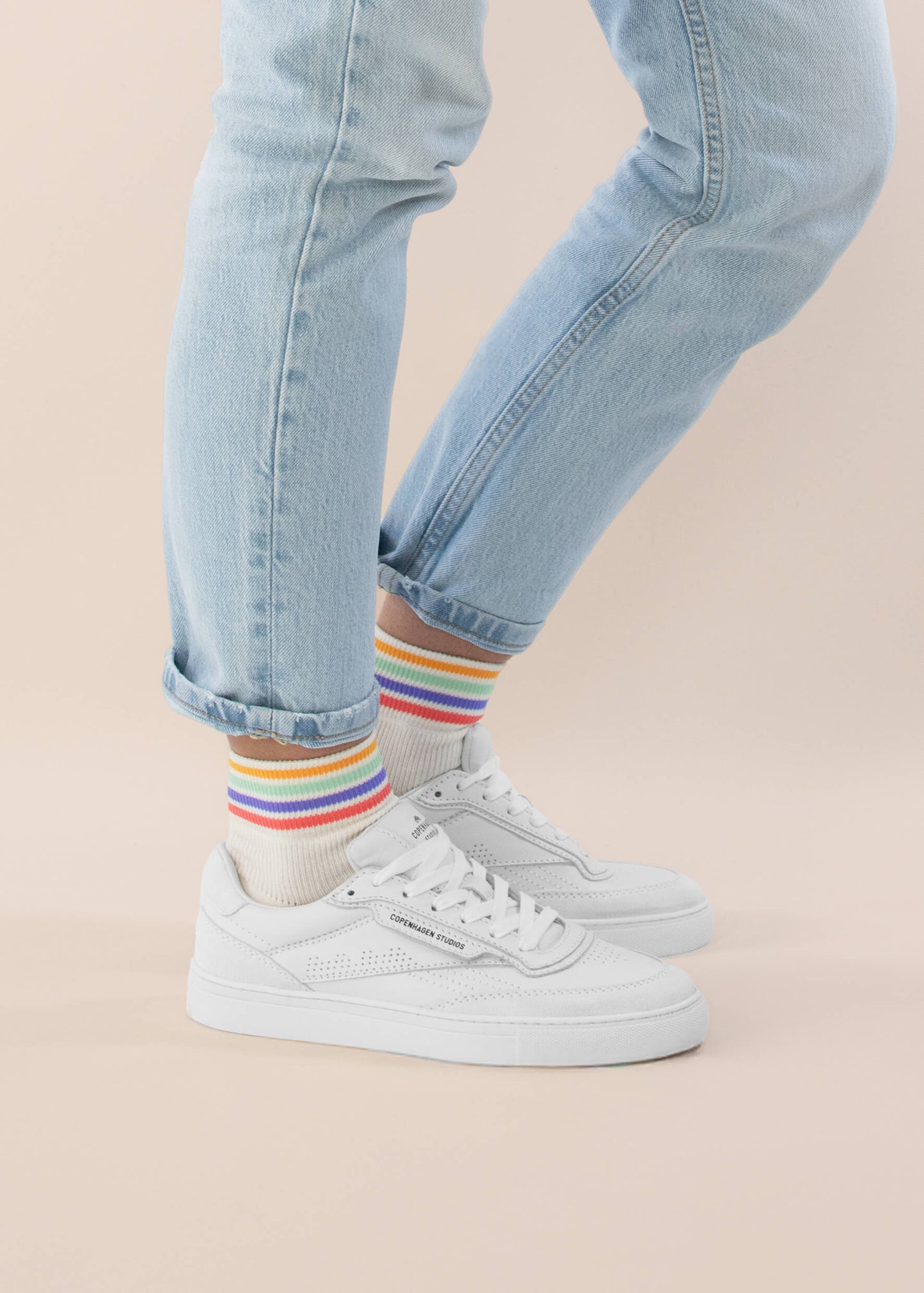 Eine Person trägt hellblaue Jeans, J.Clay Sneaker Socken aus atmungsaktivem Material mit farbigen Streifen und weiße Turnschuhe und steht mit einem Fuß leicht erhöht vor einem hellbeigen Hintergrund.