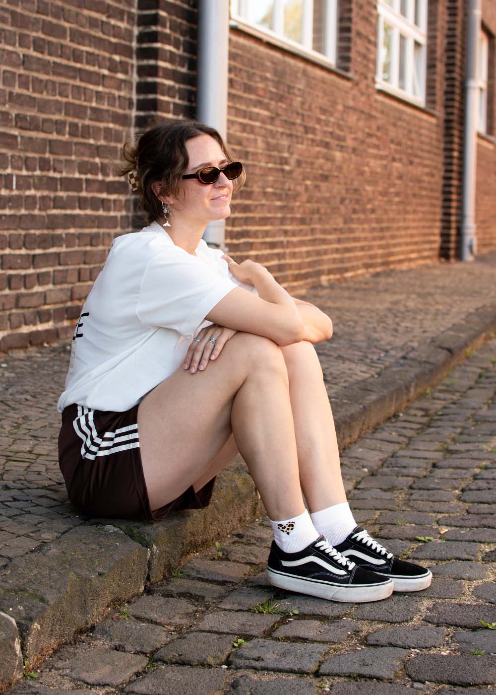 Eine Frau mit Sonnenbrille sitzt auf einem Bordstein und trägt ein „J.Clay Leo Heart – SSH“-Shirt, Shorts und Turnschuhe.