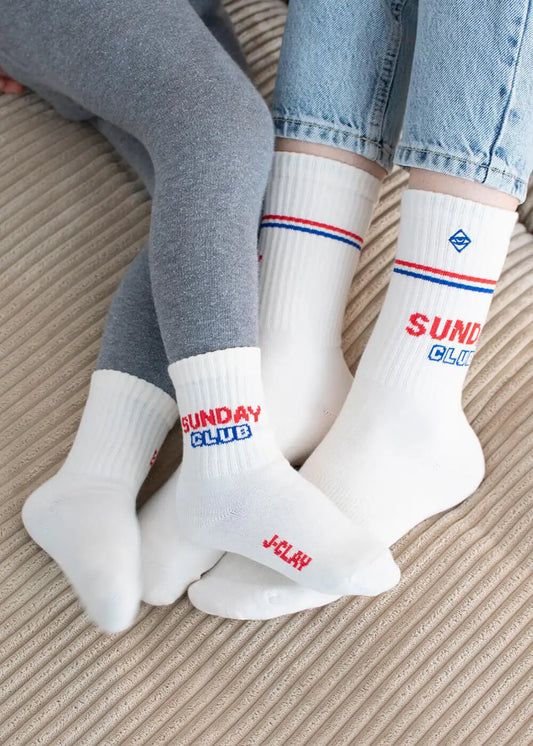 Zwei Personen sitzen eng beieinander auf einer gerippten Fläche und tragen J.Clay Partner-Look Socken mit dem SUNDAY CLUB Schriftzug in Rot und Blau. Die eine hat graue Leggings an, die andere blaue Jeans - ihre passenden Socken berühren sich, während sie nebeneinander entspannen.
