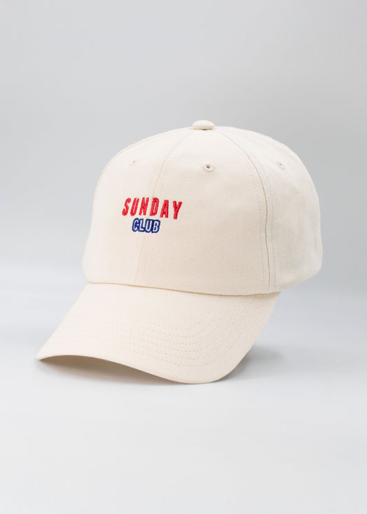 Eine beigefarbene J.Clay Dad Cap mit aufgesticktem SUNDAY CLUB in Rot und Blau auf der Vorderseite vor einem schlichten hellgrauen Hintergrund.