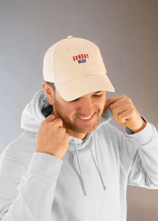 Ein lächelnder Mann trägt einen hellgrauen J.Clay-Kapuzenpulli und eine beigefarbene Vatermütze mit der Aufschrift SUNDAY CLUB in Rot und Blau, während er die Schnüre seines Kapuzenpullis vor einem neutralen Hintergrund zurechtrückt.