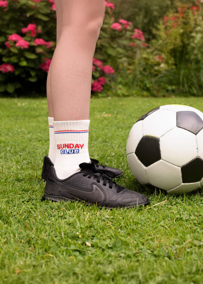 Kind in J.Clay Sunday Club - Kindersocken und schwarzen Turnschuhen steht neben einem Fußball auf dem Rasen.