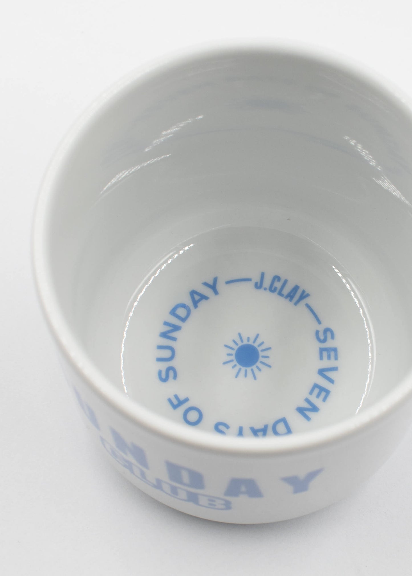 Eine weiße J.Clay Tasse ohne Henkel aus Keramik, von oben gesehen, mit blauem Text auf der Innenseite: "J.Clay-Seven Days of Sunday" um ein Sonnensymbol herum; blauer Text ist auch teilweise auf dem äußeren Rand sichtbar.