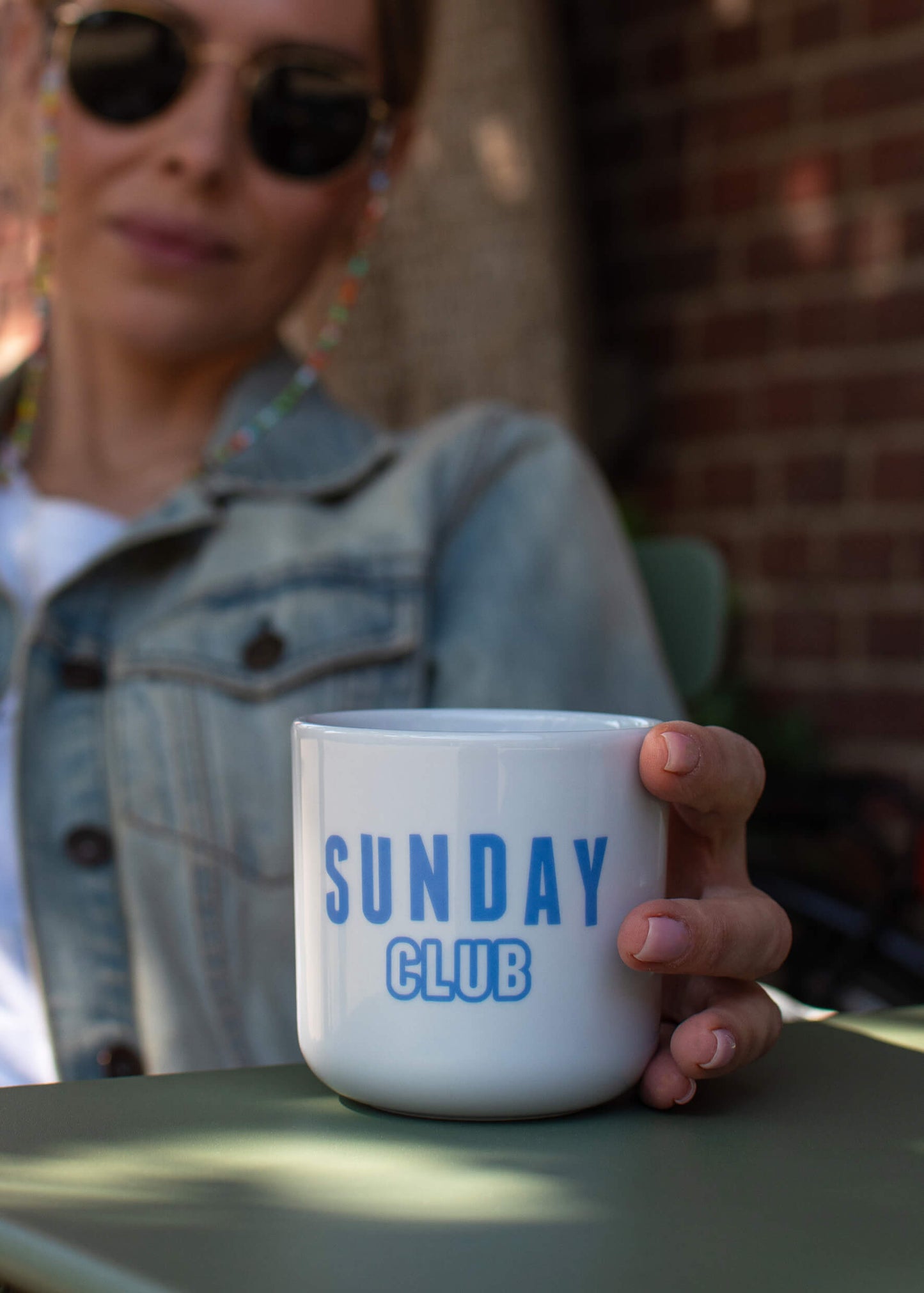 Eine Person mit Sonnenbrille und Jeansjacke hält eine J.Clay Tasse ohne Henkel mit der Aufschrift "SUNDAY CLUB" in Blau und sitzt draußen an einer Backsteinmauer. J.Clay Tennissocken lugen unter ihren Jeans hervor und tragen zur gemütlichen Geschenkset-Atmosphäre bei.