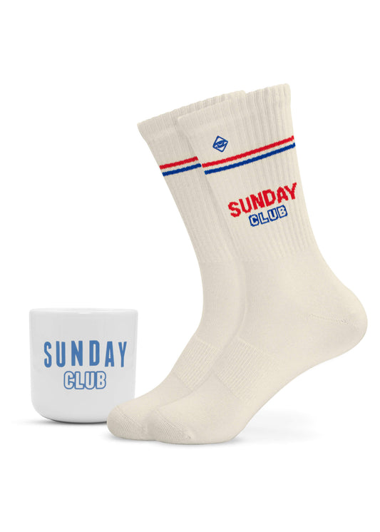 Ein Paar cremefarbene J.Clay-Tennissocken mit roten und blauen Streifen und der Aufschrift "SUNDAY CLUB", daneben ein henkelloser J.Clay-Becher mit der Aufschrift "SUNDAY CLUB" in Blau, beide auf weißem Hintergrund.