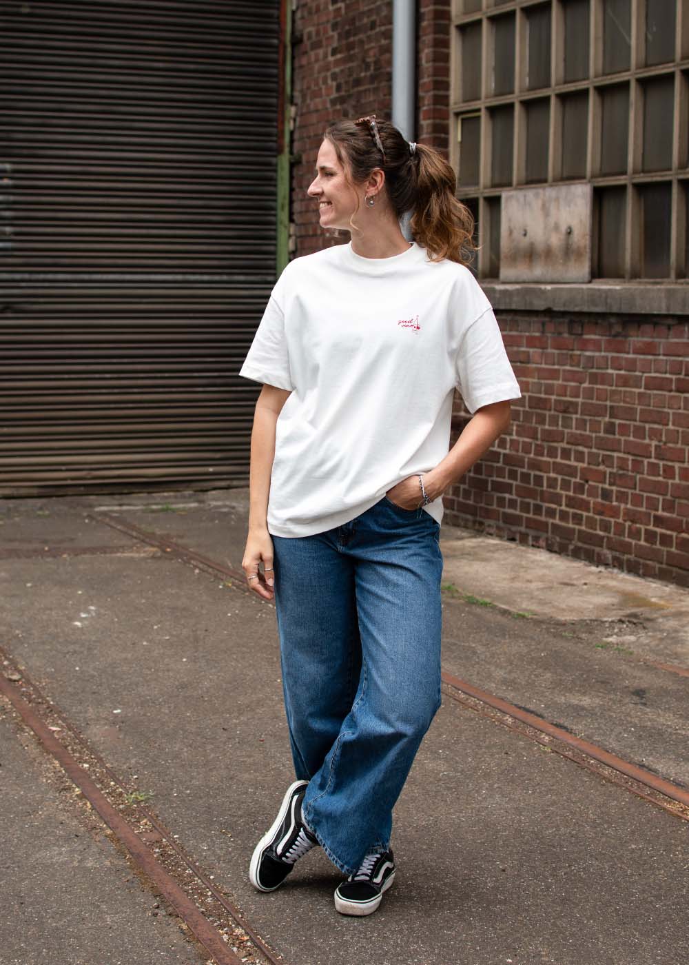 Eine Frau in einem „J.Clay Good Vino“-T-Shirt und blauen Jeans steht vor einem Backsteingebäude und schaut zur Seite.
