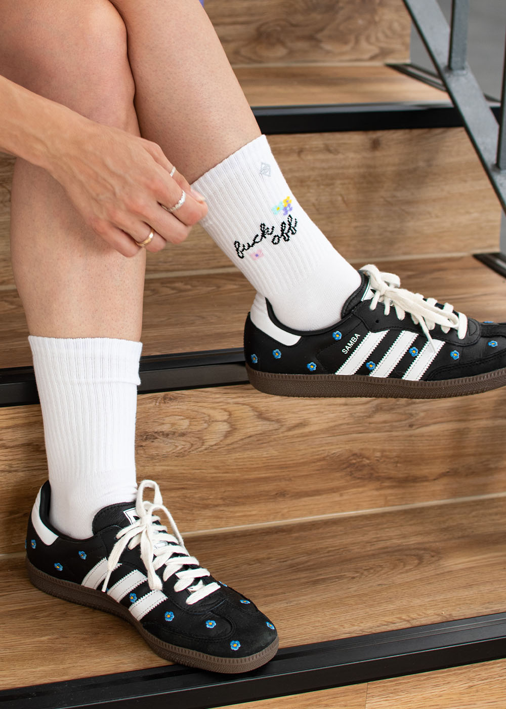 Die Person trägt schwarze Adidas-Sneaker und J.Clay „Fuck Off“-Tennissocken und sitzt auf einer Treppe.