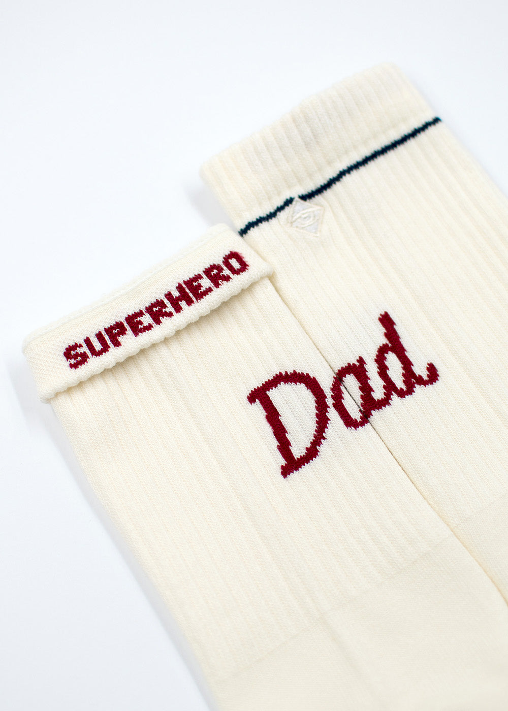 Zwei cremefarbene Socken von J.Clay: auf einem steht SUPERHERO, auf dem anderen Dad in roten Buchstaben.