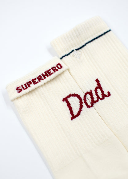Zwei cremefarbene Socken von J.Clay: auf einem steht SUPERHERO, auf dem anderen Dad in roten Buchstaben.