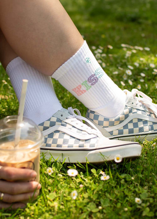 Person in karierten J.Clay Kiss Turnschuhen und hochwertigen Socken, mit Eiskaffee in der Hand auf einer Wiese.