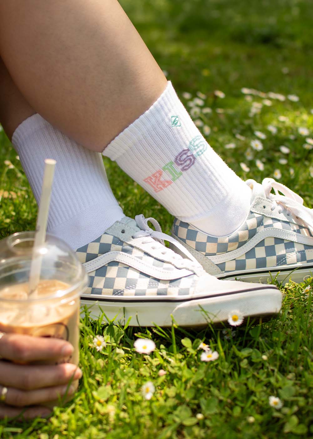 Person in karierten J.Clay Kiss Turnschuhen und hochwertigen Socken, mit Eiskaffee in der Hand auf einer Wiese.