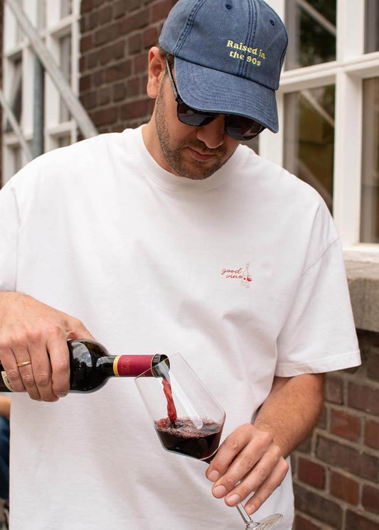 Ein Mann in einem weißen „J.Clay Good Vino“-T-Shirt schenkt Rotwein ein; er trägt eine blaue Mütze und eine Sonnenbrille.
