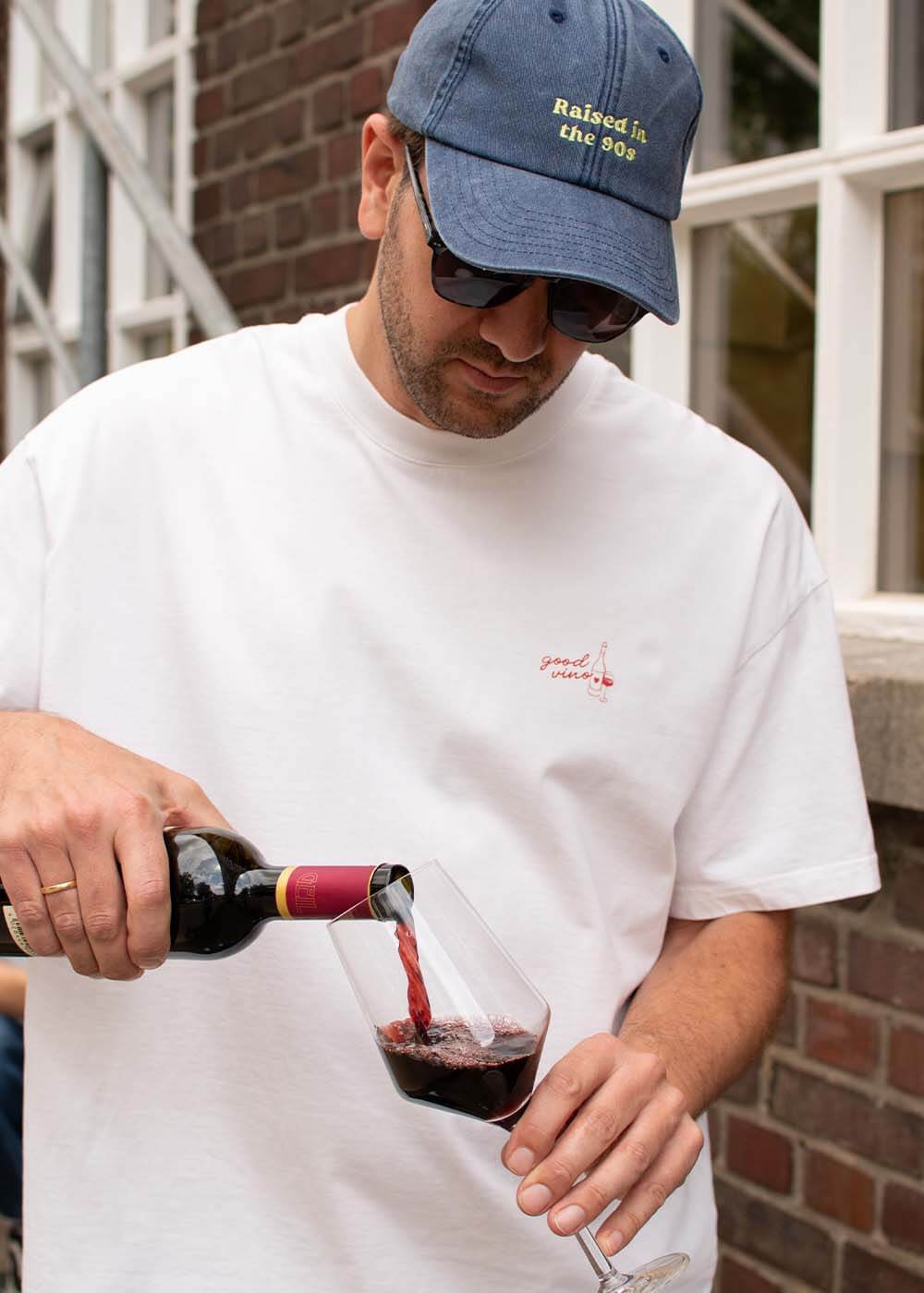 Ein Mann in einem weißen „J.Clay Good Vino“-T-Shirt schenkt Rotwein ein; er trägt eine blaue Mütze und eine Sonnenbrille.