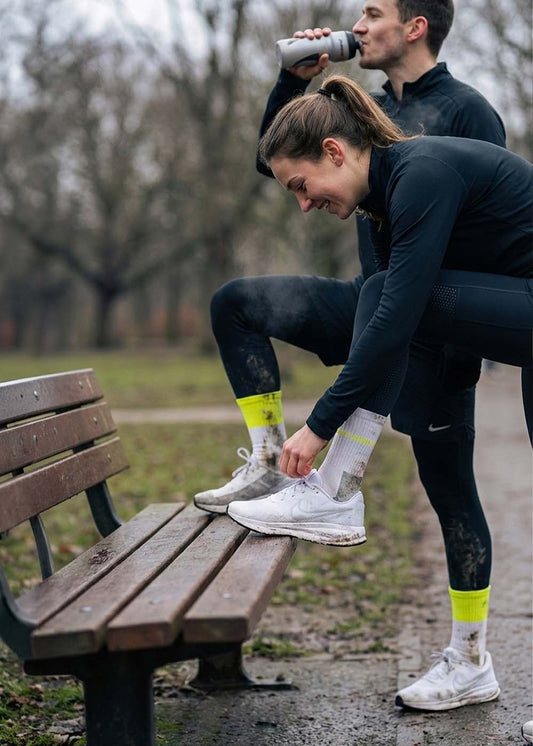 Zwei in Sportkleidung - einer bindet J.Clay Neon Block Sportsocken an, einer trinkt Wasser im Freien.