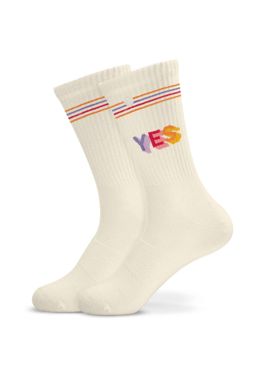 Ein Paar cremefarbene Tennissocken von J.Clay mit Regenbogenstreifen an der Oberseite und dem Wort "YES" in bunten Buchstaben an der Seite, die vor einem weißen Hintergrund stehen.