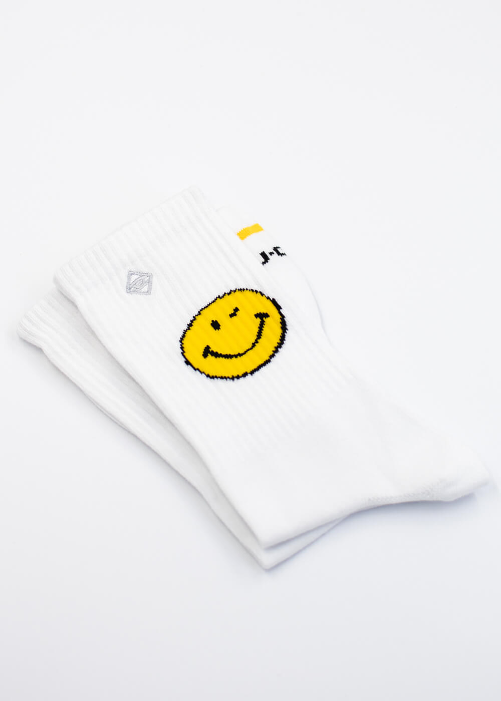 Ein Paar weiße Tennissocken von J.Clay Yellow Smile, entworfen in Deutschland, mit einer großen gelben Smiley-Grafik an der Seite und ordentlich gefaltet auf weißem Hintergrund.