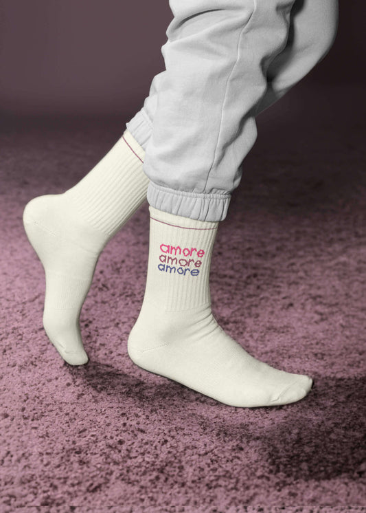 Eine Person in hellgrauer Jogginghose steht auf Zehenspitzen auf einem lilafarbenen Strukturteppich und trägt weiße J.Clay-Tennissocken, auf denen das Wort "amore" dreimal in Rosa und Lila aufgedruckt ist.