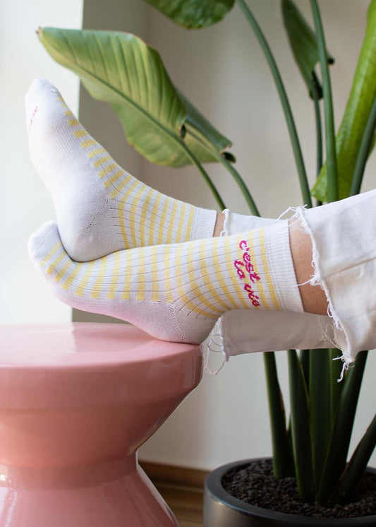 Die Füße in „C’est la vie – Midi“ von J.Clay ruhen auf einem rosa Hocker, im Hintergrund sind grüne Pflanzen zu sehen.