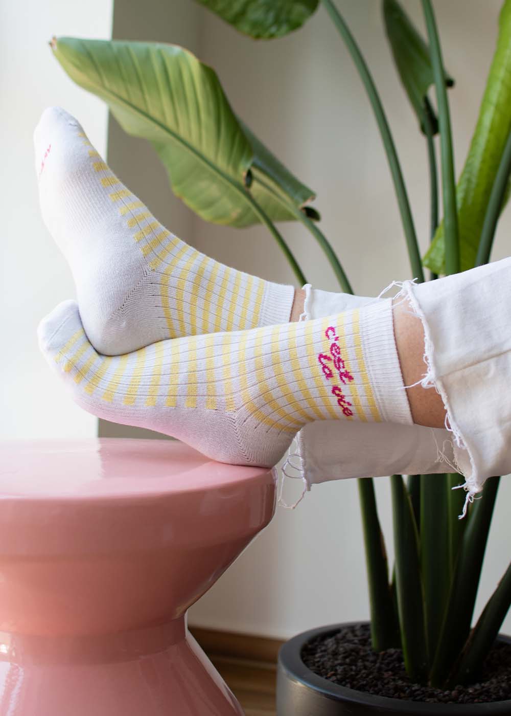 Die Füße in „C’est la vie – Midi“ von J.Clay ruhen auf einem rosa Hocker, im Hintergrund sind grüne Pflanzen zu sehen.