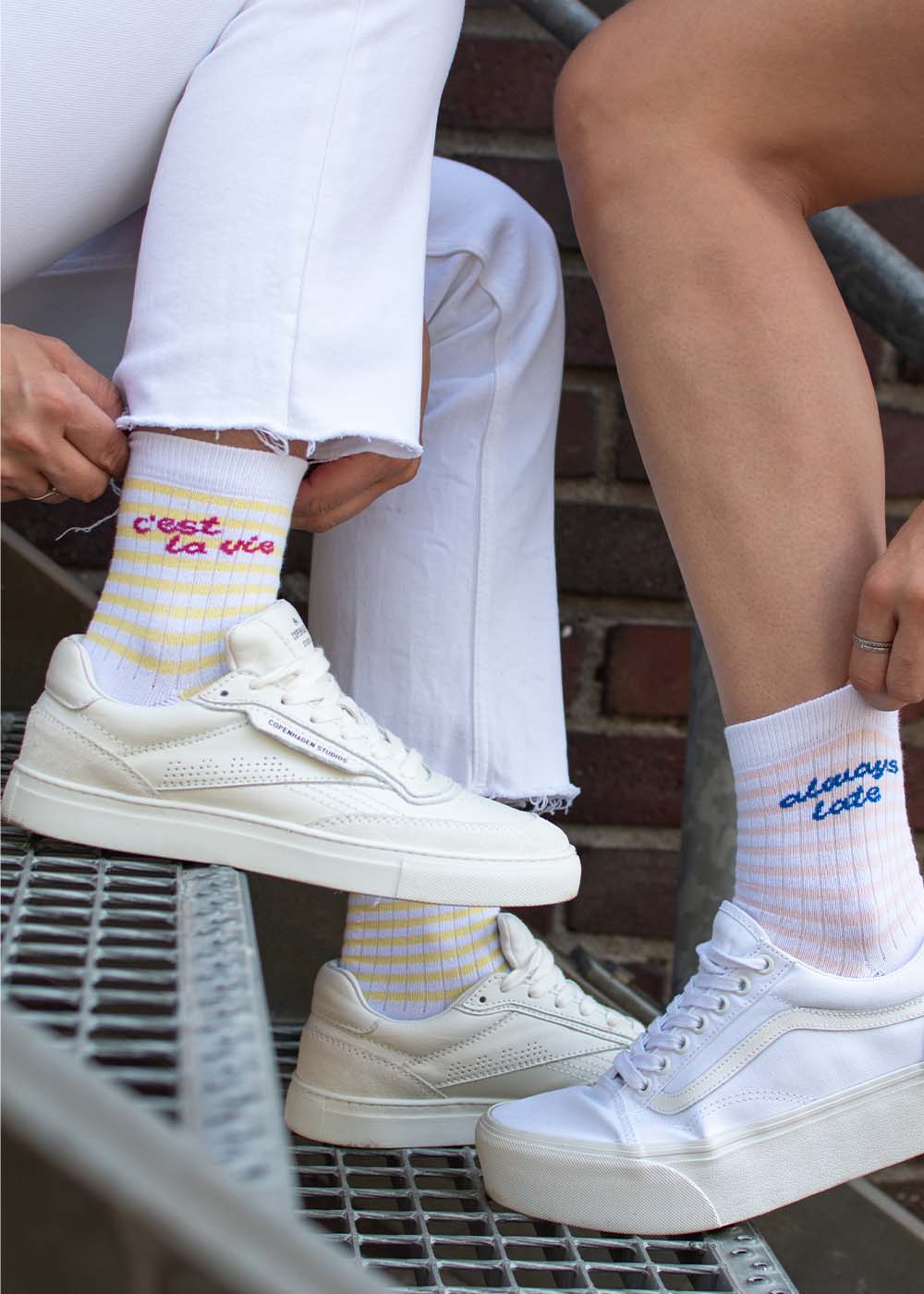 Zwei Personen in weißen Hosen und Sneakers präsentieren die „C’est la vie – Midi Pastell“-Socken von J.Clay.