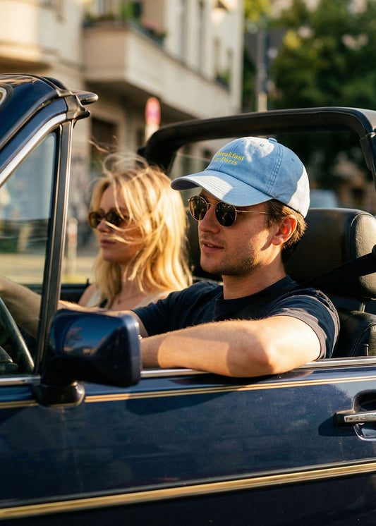 Zwei Personen mit Sonnenbrillen und einer J.Clay Breakfast in Paris Cap sitzen in einem blauen Cabrio.