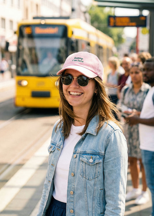 Lächelnde Frau mit einer J.Clay "Sorry I'm late"-Mütze und Sonnenbrille an einer Straßenbahnhaltestelle mit einer gelben Straßenbahn.
