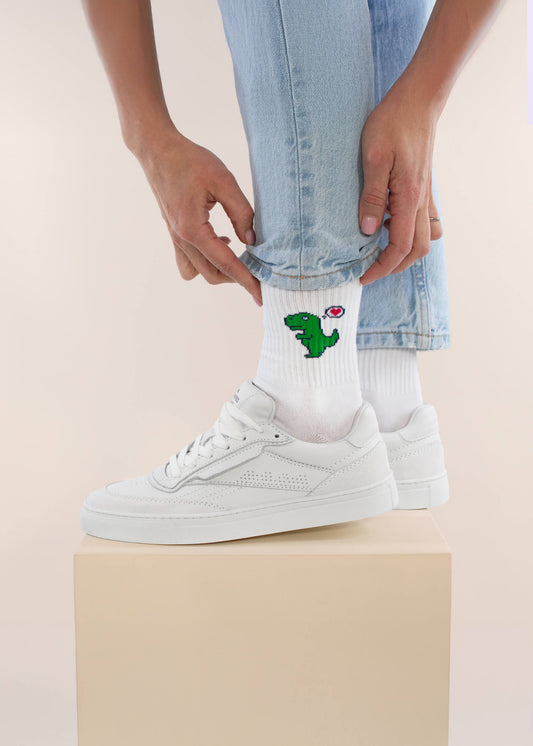 Eine Person in hellblauen Jeans und weißen Turnschuhen krempelt ihr Hosenbein hoch und zeigt die trendigen J.Clay Tennissocken mit einem grünen Dinosaurier und einem roten Herz, die auf einer beigen Schachtel stehen.