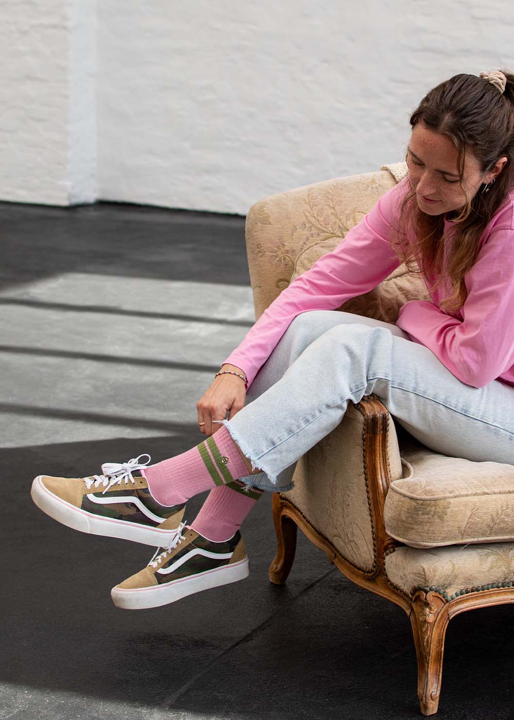 Eine Frau in einem rosa Oberteil und J.Clay True Rose Olive Tennissocken sitzt lächelnd auf einem beigen Stuhl.