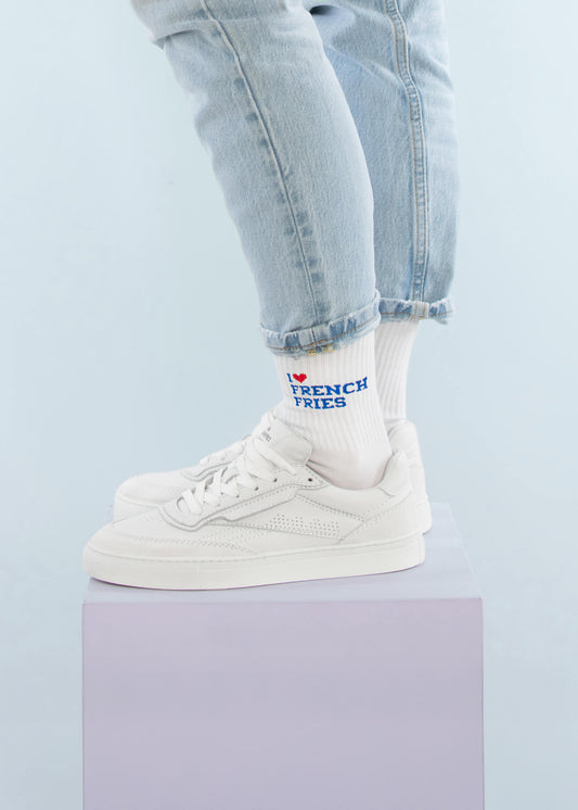Eine Person in hellblauen Jeans mit Bündchen, weißen Turnschuhen und J.Clay-Tennissocken mit dem Schriftzug "I ❤️ FRENCH FRIES" steht auf einem pastellfarbenen lila Würfel vor hellblauem Hintergrund.