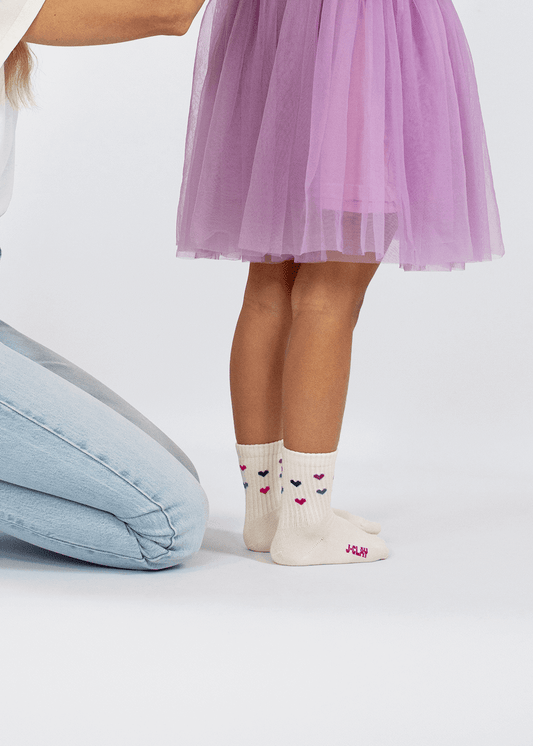Drei Paar cremefarbene J.Clay-Babysocken (0-4 Jahre) mit Rippbündchen, kleinen rosa, lila und schwarzen Herzen und dem Aufdruck "JCLAY" in rosa auf der Sohle. - Mini Socken 0-4 Jahre