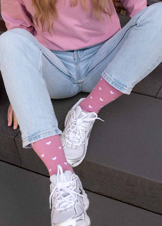 Eine Person in Jeans und J.Clay-Socken mit „Hearts“-Motiv, die auf dunklen Stufen sitzt und weiße Turnschuhe trägt.