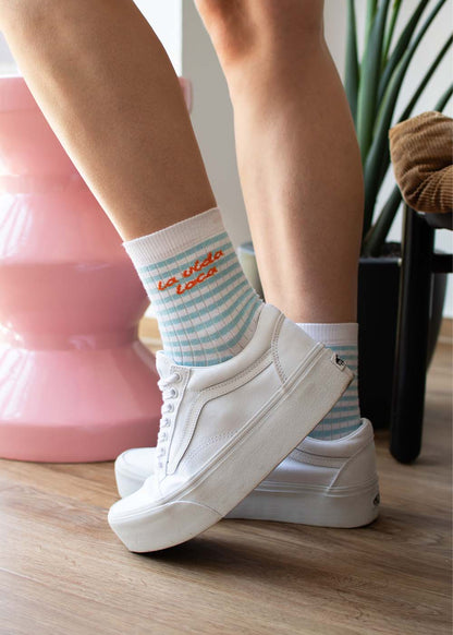 Person, die J.Clay „la vida loca“ – blau karierte Midi-Socken trägt und in weißen Sneakers in einem Innenraum steht.