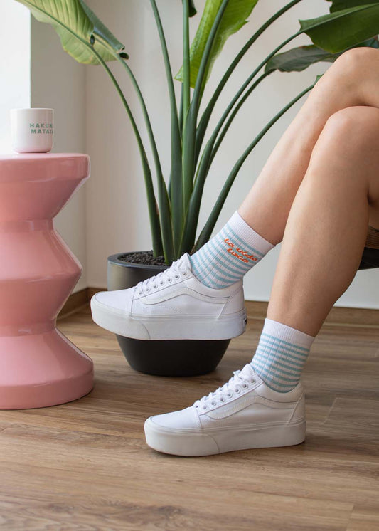 Eine Person trägt „J.Clay la vida loca“ – weiße Midi-Sneaker und gestreifte Socken und steht neben Pflanzen auf einem Holzboden.
