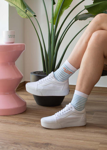 Eine Person trägt „J.Clay la vida loca“ – weiße Midi-Sneaker und gestreifte Socken und steht neben Pflanzen auf einem Holzboden.