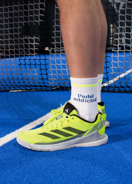 Ein Paar weiße J.Clay Sportsocken mit gelben Streifen und "Padel addicted" in Blau - ideal für Padel-Tennis-Fans. Der Hintergrund ist weiß mit einem blauen X SPORTS Logo in der oberen rechten Ecke.