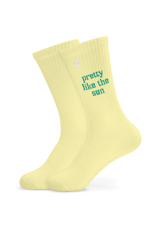 Ein Paar hellgelbe Tennissocken von J.Clay mit der Aufschrift "pretty like the sun" in Petrol auf der Seite, die sich von einem weißen Hintergrund abhebt.