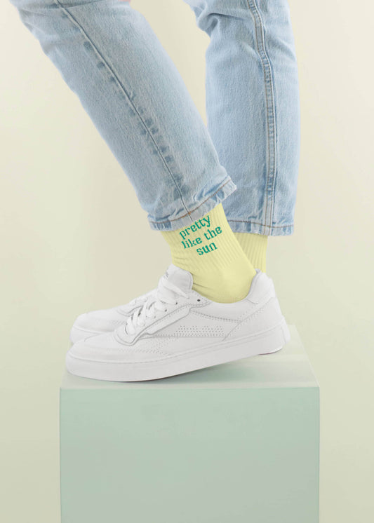 Eine Person steht auf einem hellpastellgrünen Podest vor einem hellen Hintergrund, trägt eine hellblaue Jeans von J.Clay, gelbe Tennissocken mit "pretty like the sun" und weiße Turnschuhe.