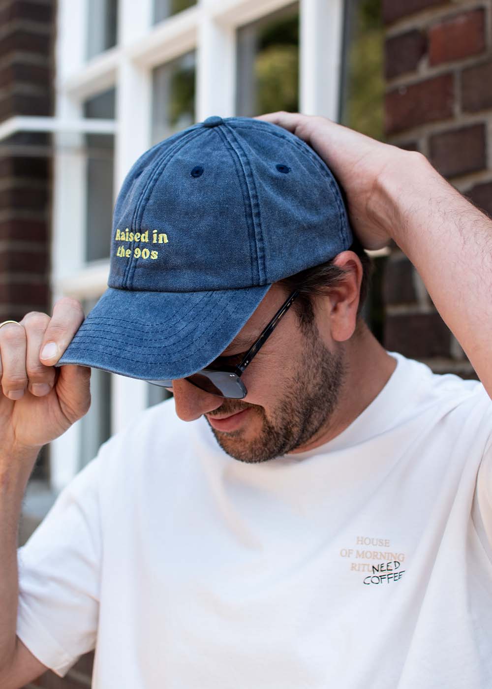 Ein Mann mit einer blauen Kappe von J.Clay Raised in the 90s, Sonnenbrille und weißem Hemd, der seine Kappe zurechtzieht.