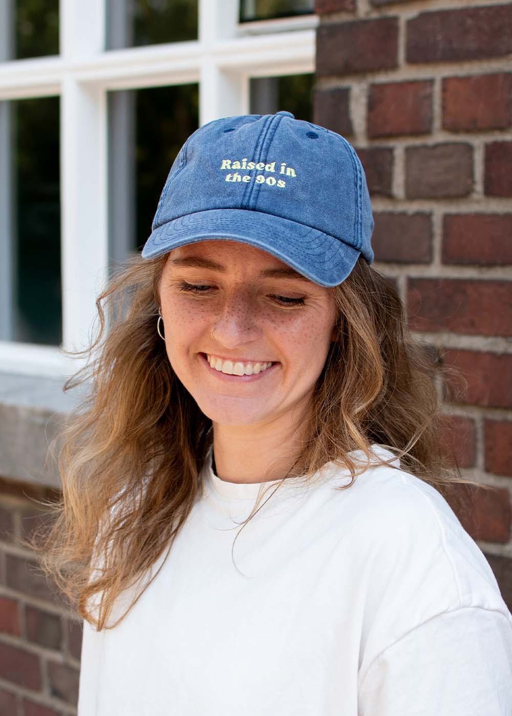 Eine lächelnde Person mit einer blauen „J.Clay Raised in the 90s“-Kappe und einem weißen Hemd vor einer Backsteinmauer.