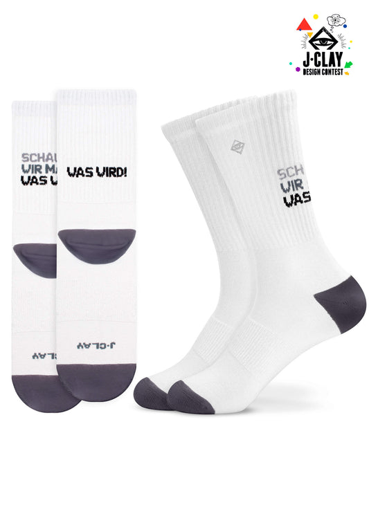 Zwei Paar weiße J.Clay Tennissocken mit grauen Fersen und Zehen. Auf der linken Socke steht "SCHAUEN WIR MAL, WAS WIRD!", auf der rechten "SCHAUEN WIR MAL, WAS." Schwarzer deutscher Text; J.Clay Design Contest Logo oben rechts.