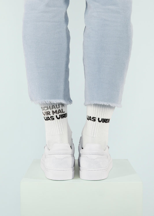 Eine Person, die eine hellblaue Cordhose von J.Clay, Tennissocken mit schwarzem Text und weiße Turnschuhe trägt, steht auf einem weißen Podest vor einem hellen Hintergrund; nur die Unterschenkel und Füße sind sichtbar.