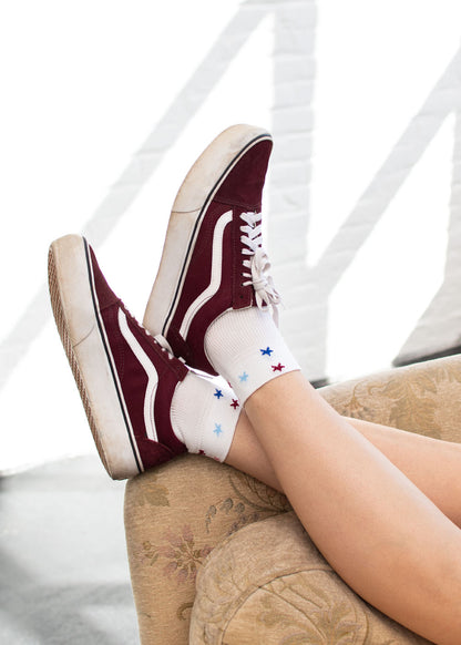 Eine Person trägt Socken von J.Clay Stars – SSH und kastanienbraune Turnschuhe; ihre Füße ruhen auf einem beigen Sofa.
