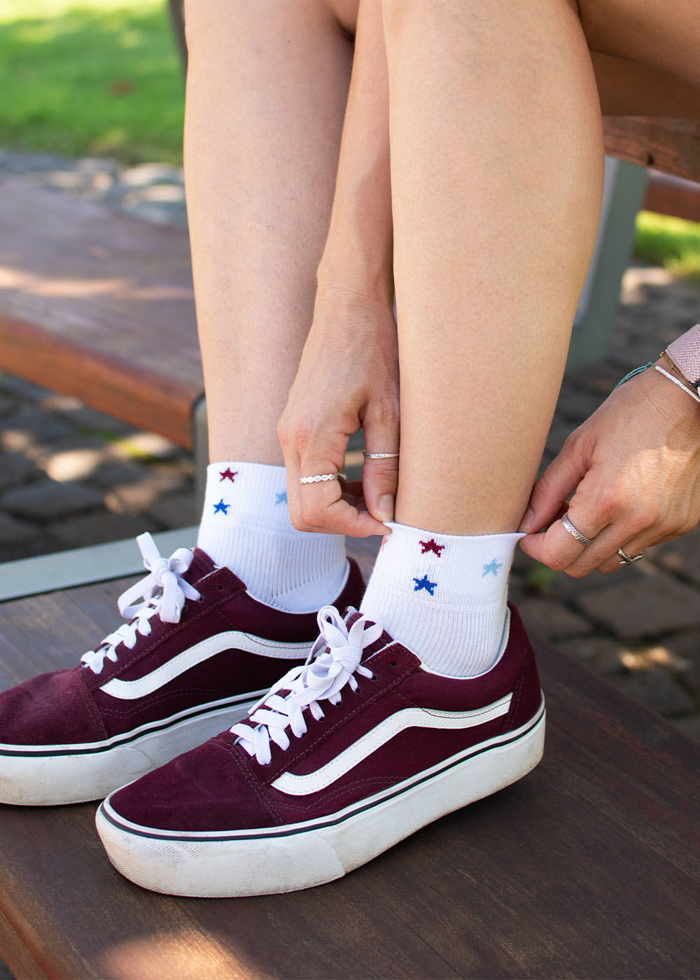 Auf einer Holzbank sitzen Hände, die J.Clay Stars – SSH-Socken – mit kastanienbraunen Sneakers zurechtziehen.